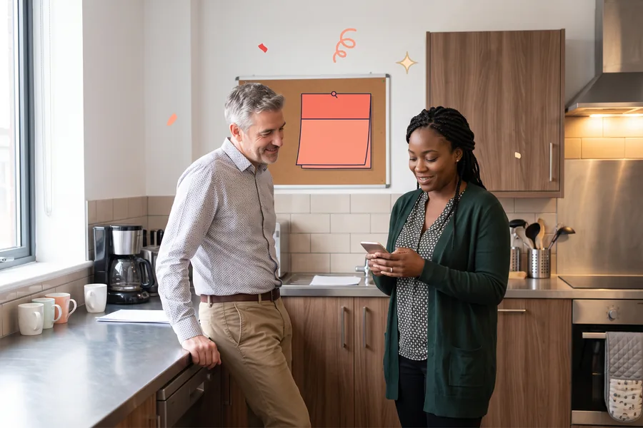 Two coworkers in an office kitchen smiling as she reads her phone