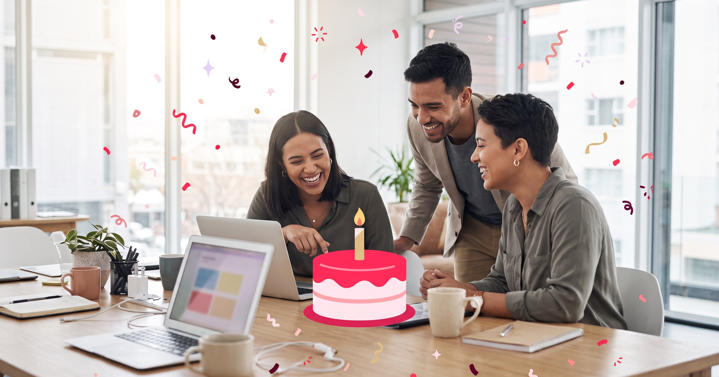 Coworkers laughing together at a desk as a teammate reads a birthday message on her laptop