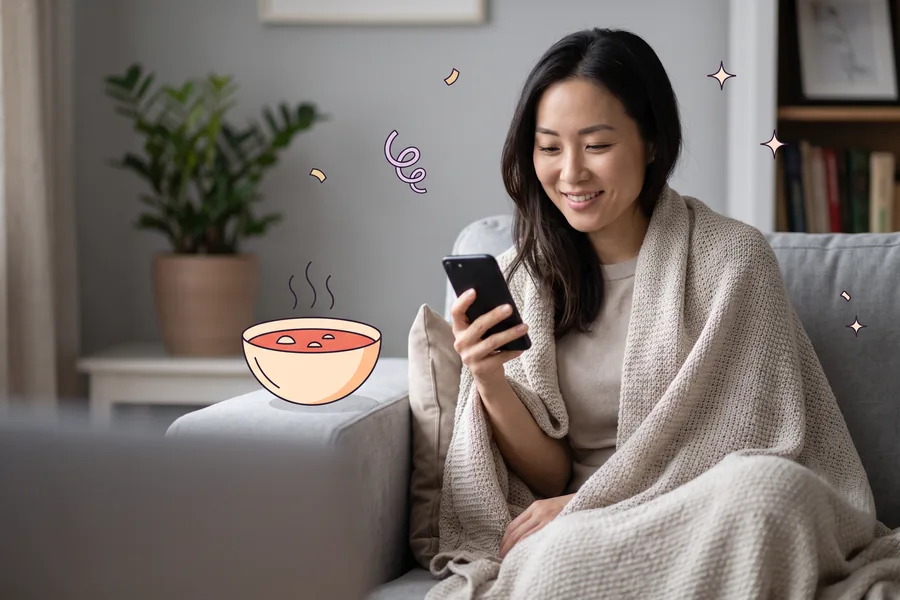 Woman wrapped in a blanket on her sofa, smiling at her phone, a bowl of soup beside her